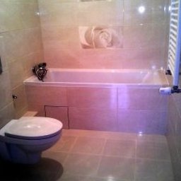 Flismarbud - Beżowa łazienka z wanną obłożoną płytkami, nad wanną dwa kwadratowe kafelki z motywem kwiatowym, toaleta w rogu, grzejnik drabinkowy po prawej.