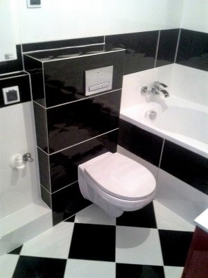 Nowoczesna łazienka w Szczecinie z czarno-białymi płytkami ułożonymi w szachownicę na podłodze, wisząca toaleta z zabudową geberit wykończoną czarnymi płytkami, biała wanna z baterią.