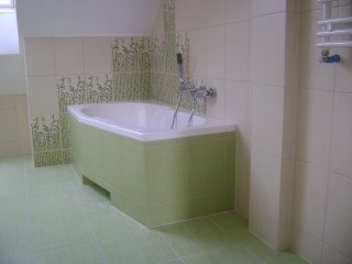 Biała wanna obudowana zielonymi płytkami w łazience z beżowymi i zielonymi płytkami ściennymi z motywem roślinnym, widok z boku.