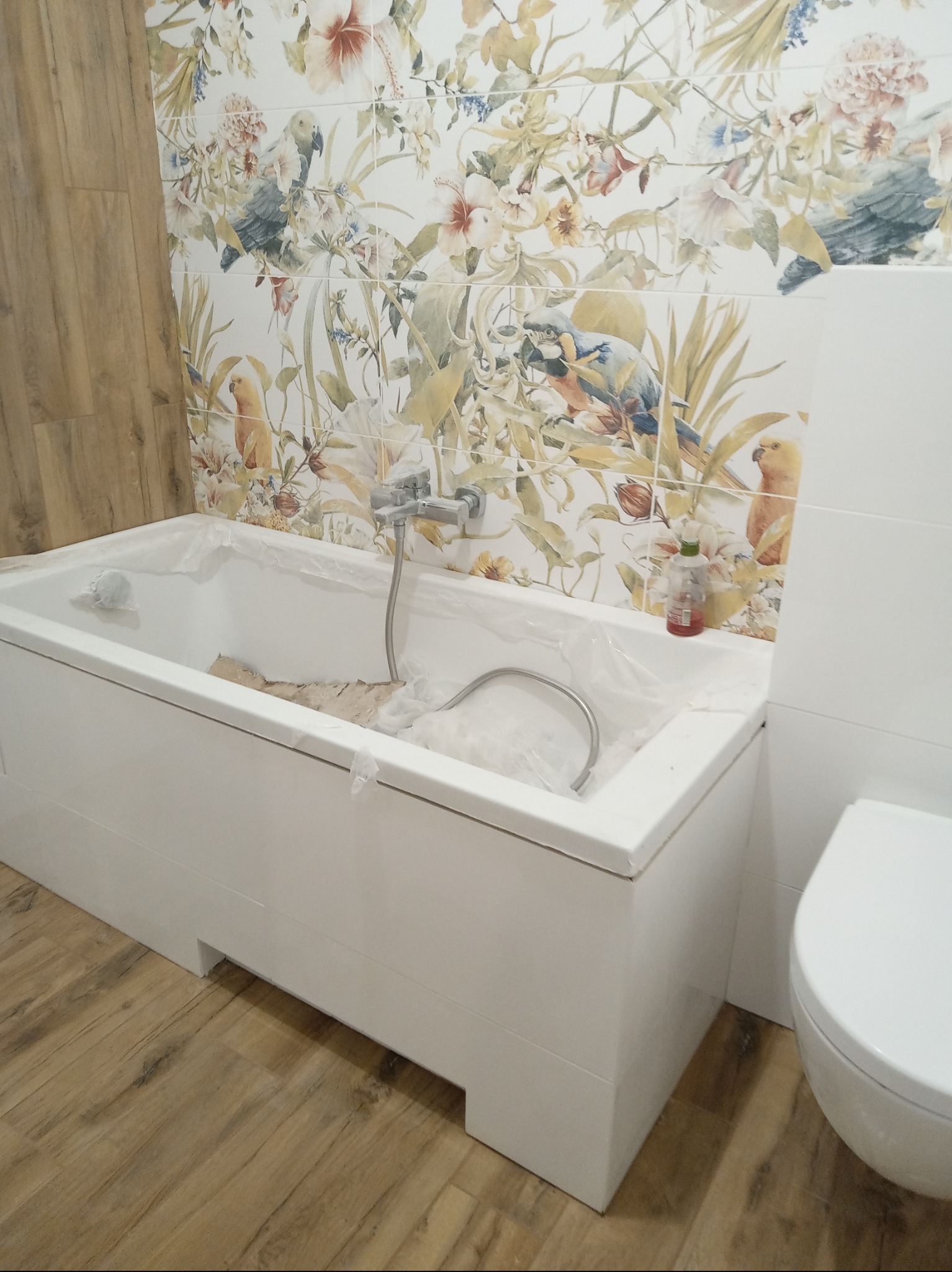 Biała wanna obłożona folią ochronną podczas prac remontowych, ściany wyłożone płytkami z motywem kwiatowym i ptasim, drewnopodobna podłoga, widoczna bateria wannowa.