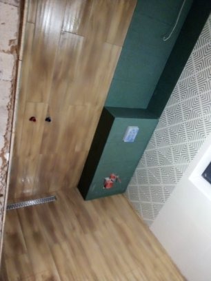 Fragment łazienki w trakcie remontu: drewnopodobne płytki na podłodze, zielony element zabudowy z instalacją, ściana z wzorzystą tapetą, widoczny odpływ liniowy.