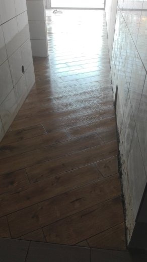Ułożona pod kątem brązowa podłoga z płytek imitujących drewno w wąskim korytarzu łazienki, białe płytki na ścianach w trakcie montażu, widoczne fugi i niedokończone wykończenia.