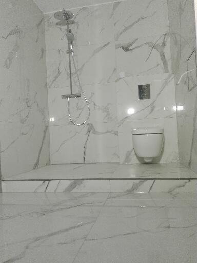 Wykończona łazienka z białą armaturą i płytkami imitującymi marmur, widoczny prysznic i podwieszana toaleta, całość utrzymana w jasnej kolorystyce.