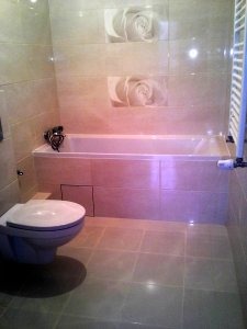 Beżowa łazienka z wanną obłożoną płytkami, nad wanną dwa kwadratowe kafelki z motywem kwiatowym, toaleta w rogu, grzejnik drabinkowy po prawej.