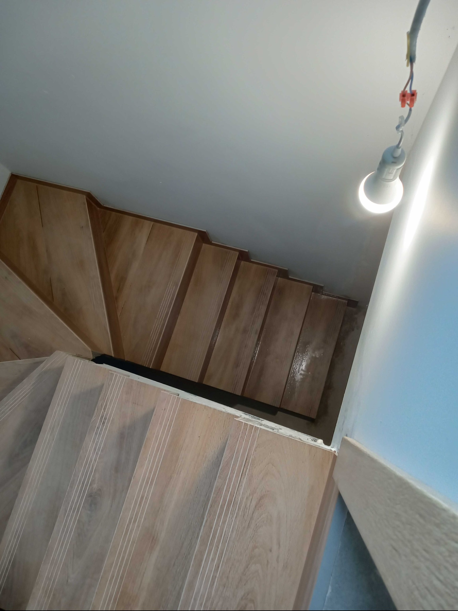 Widok z góry na drewniane schody z białymi intarsjami na stopniach, oświetlone wiszącą żarówką na kablu, z widocznymi złączkami elektrycznymi.