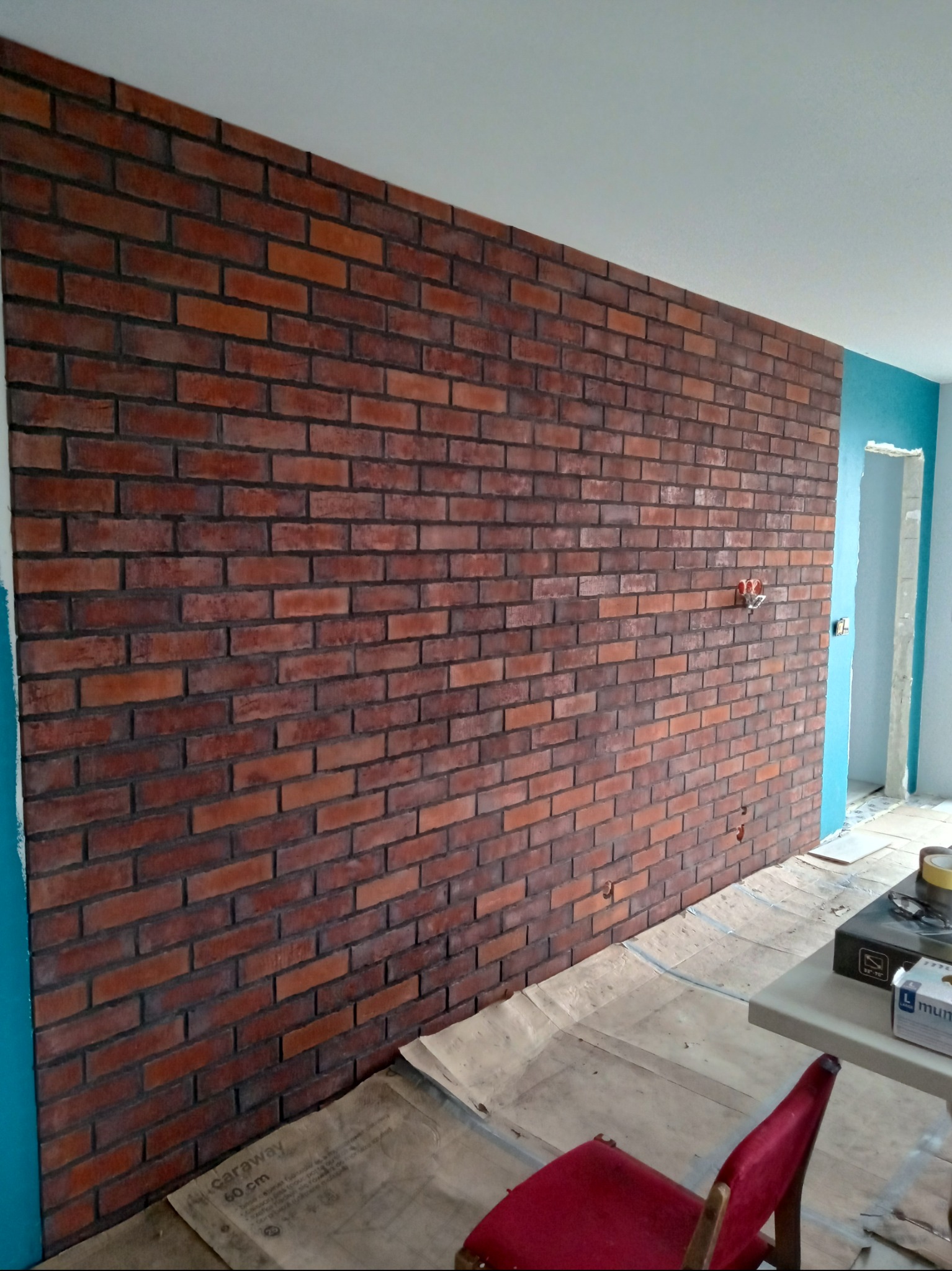 Ściana wykończona czerwoną cegłą klinkierową z ciemną fugą. Widoczne gniazdko elektryczne i zabezpieczona folią podłoga. Czerwone krzesło w rogu kadru.