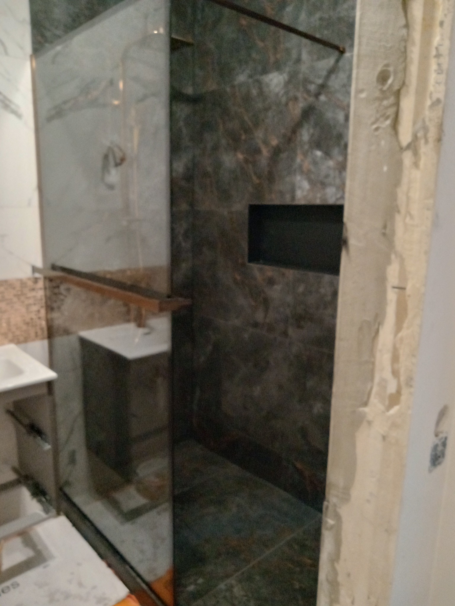 Nowoczesna kabina prysznicowa z ciemnymi płytkami imitującymi marmur, wnęka na kosmetyki, szklana ściana i widoczne prace wykończeniowe na ścianie obok.