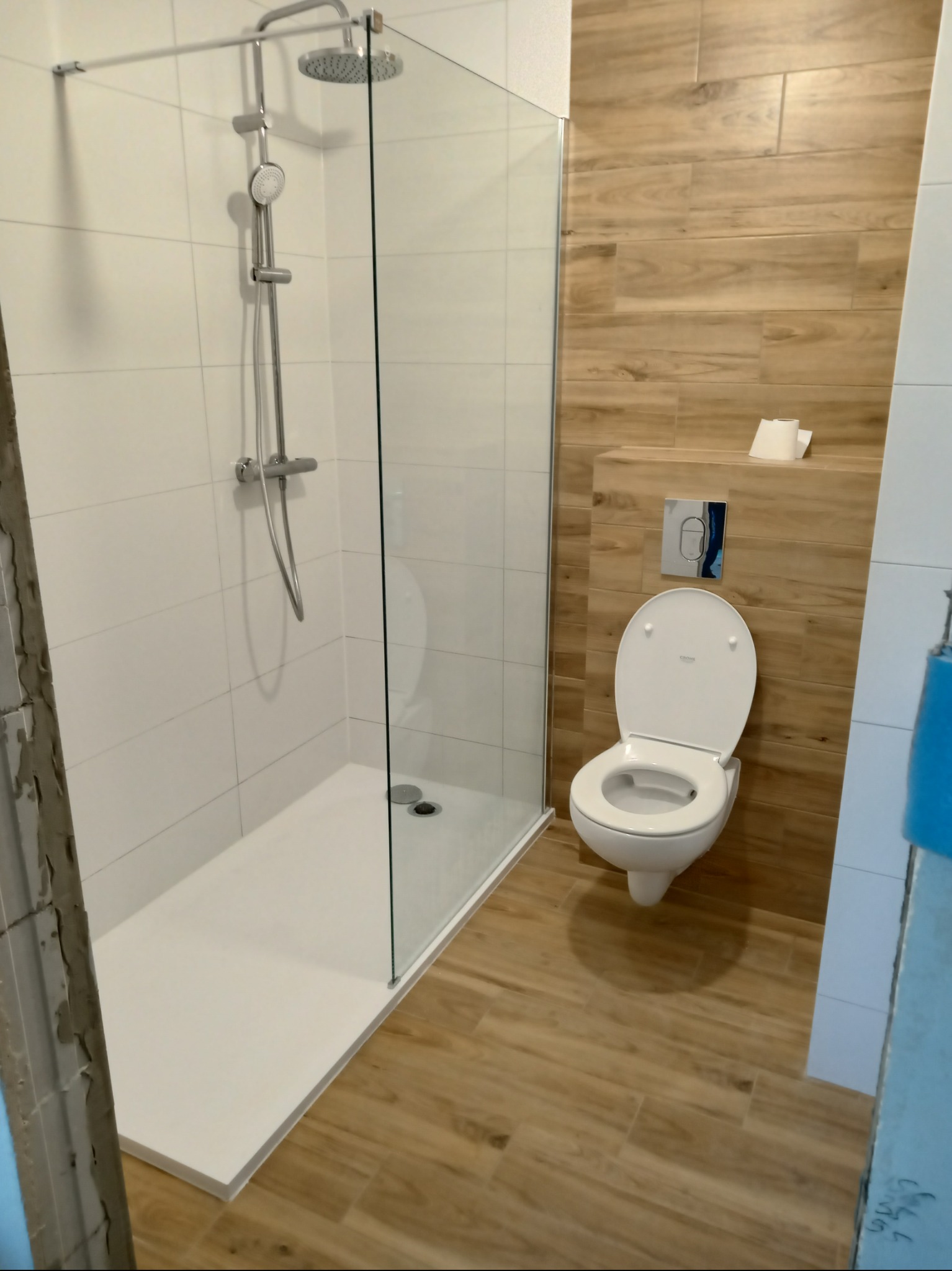 Nowoczesna łazienka po remoncie: kabina prysznicowa typu walk-in z deszczownicą, toaleta wisząca i płytki imitujące drewno na ścianie. Minimalistyczny design.