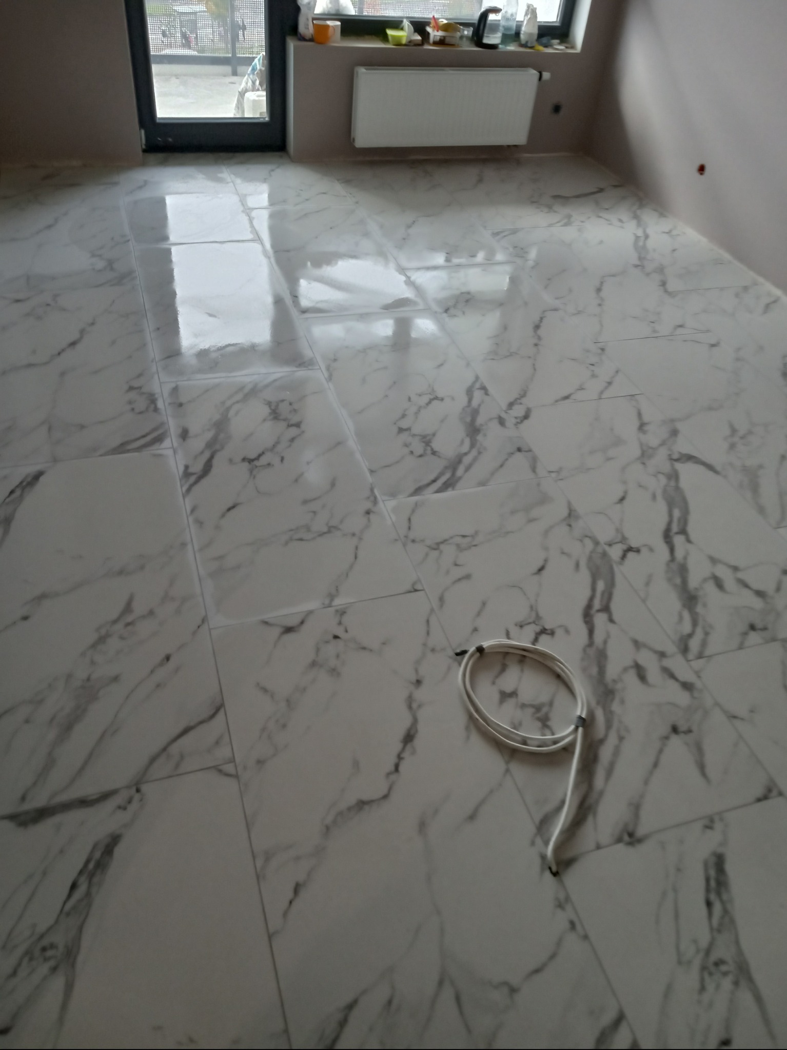Błyszcząca podłoga z płytek imitujących marmur w jasnym pomieszczeniu z oknem. Widoczny kabel na podłodze. Minimalistyczne wnętrze po remoncie.
