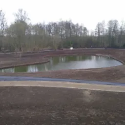 Krajobraz po pracach ziemnych z nowo wykopanym stawem o nieregularnym kształcie, otoczonym drewnianym obrzeżem i świeżo utwardzoną ścieżką, w tle drzewa bez liści.