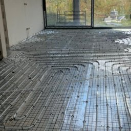 Szelbi-Instal Krzysztof Starmach - Rozłożone rury ogrzewania podłogowego na folii izolacyjnej w surowym wnętrzu z dużym oknem i widokiem na zewnątrz.