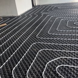 Szelbi-Instal Krzysztof Starmach - Białe rury ułożone w pętle na czarnej macie podłogowej w surowym pomieszczeniu, przygotowanie do wylewki.