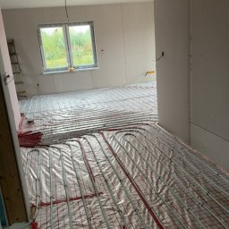 Szelbi-Instal Krzysztof Starmach - Wnętrze w trakcie montażu ogrzewania podłogowego: rury grzewcze ułożone na folii z czerwoną siatką, widok przez otwór drzwiowy, surowe ściany w tle.