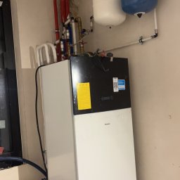 Szelbi-Instal Krzysztof Starmach - Biała pompa ciepła Daikin z podłączonymi rurami i zbiornikami wyrównawczymi w kolorze białym i niebieskim, zamontowana w pomieszczeniu z surowymi ścianami.