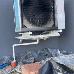 Szelbi-Instal Krzysztof Starmach - Zewnętrzna jednostka pompy ciepła zamontowana na ścianie budynku, z widocznym odprowadzeniem skroplin. W tle widoczna ściana budynku, a na pierwszym planie gruz i resztki materiałów budowlanych.