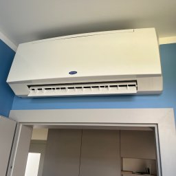 Szelbi-Instal Krzysztof Starmach - Biały klimatyzator Carrier zamontowany na błękitnej ścianie nad białymi drzwiami, widoczne żaluzje w pozycji otwartej.