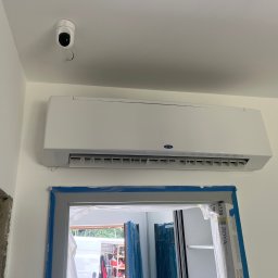 Szelbi-Instal Krzysztof Starmach - Biały klimatyzator Carrier zamontowany nad oknem w trakcie remontu, zabezpieczonym folią i taśmą malarską, z widoczną kamerą bezpieczeństwa na suficie i fragmentem dekoracyjnej lampy w rogu.