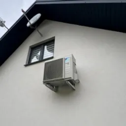 Zamontowana jednostka zewnętrzna klimatyzacji Daikin na elewacji domu z oknem i antenami satelitarnymi. Widoczne wsporniki montażowe i fragment dachu.