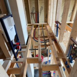 Szelbi-Instal Krzysztof Starmach - Instalacja podtynkowa WC Grohe w drewnianej konstrukcji. Widoczne rury doprowadzające wodę (czerwone i niebieskie) oraz elementy montażowe. Całość w surowym stanie, przed wykończeniem.