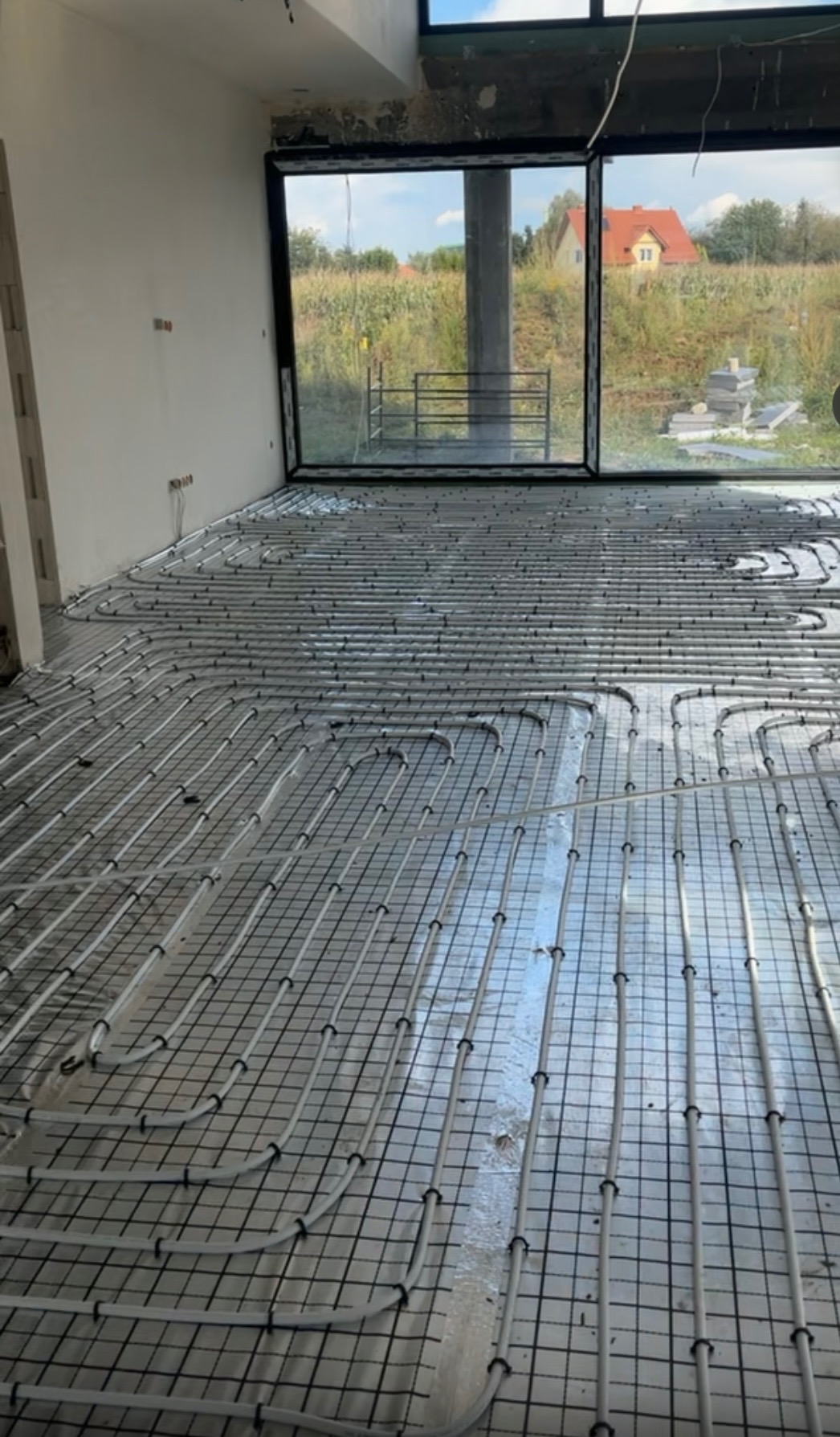 Rozłożone rury ogrzewania podłogowego na folii izolacyjnej w surowym wnętrzu z dużym oknem i widokiem na zewnątrz.