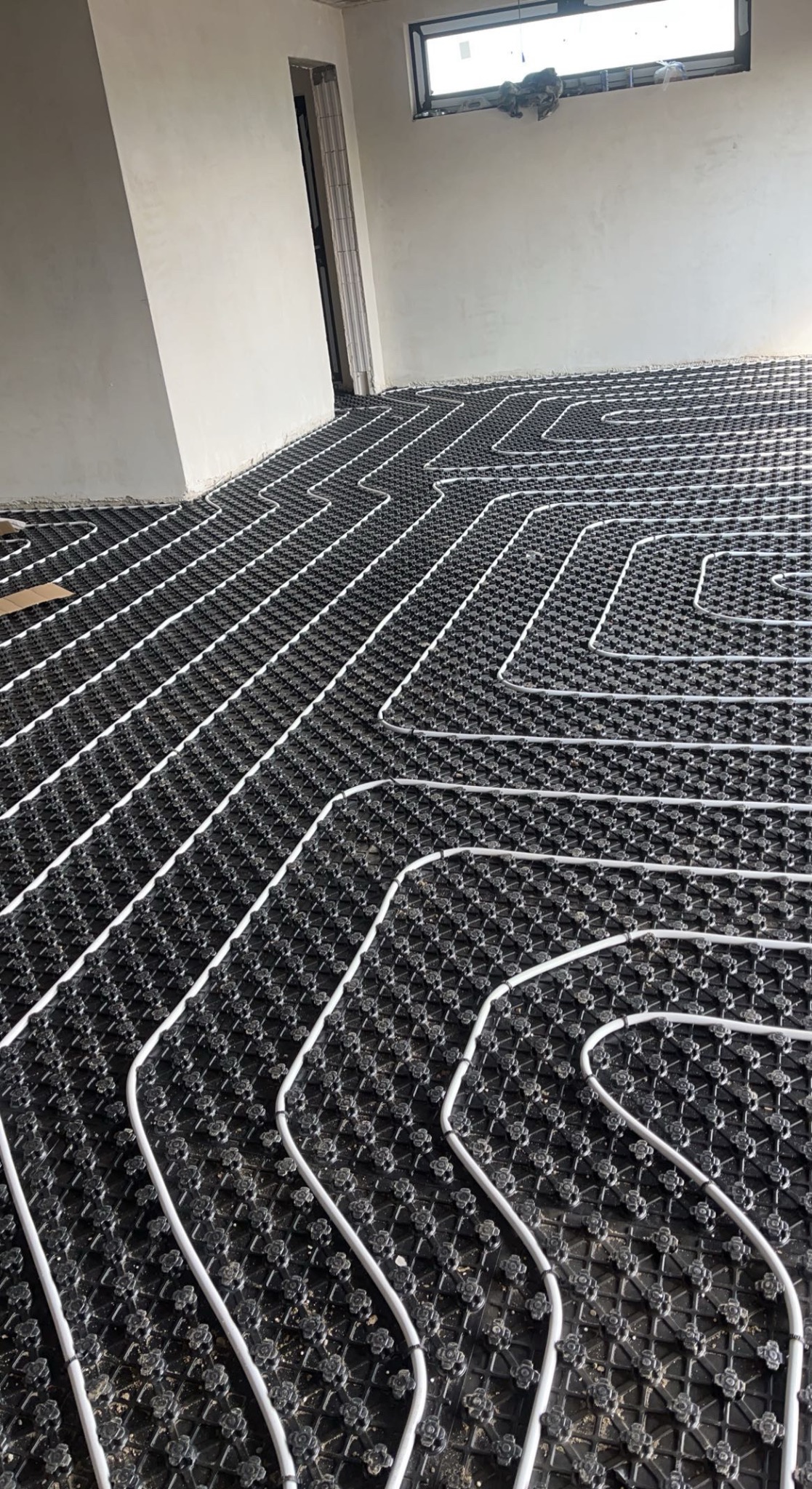 Białe rury ułożone w pętle na czarnej macie podłogowej w surowym pomieszczeniu, przygotowanie do wylewki.