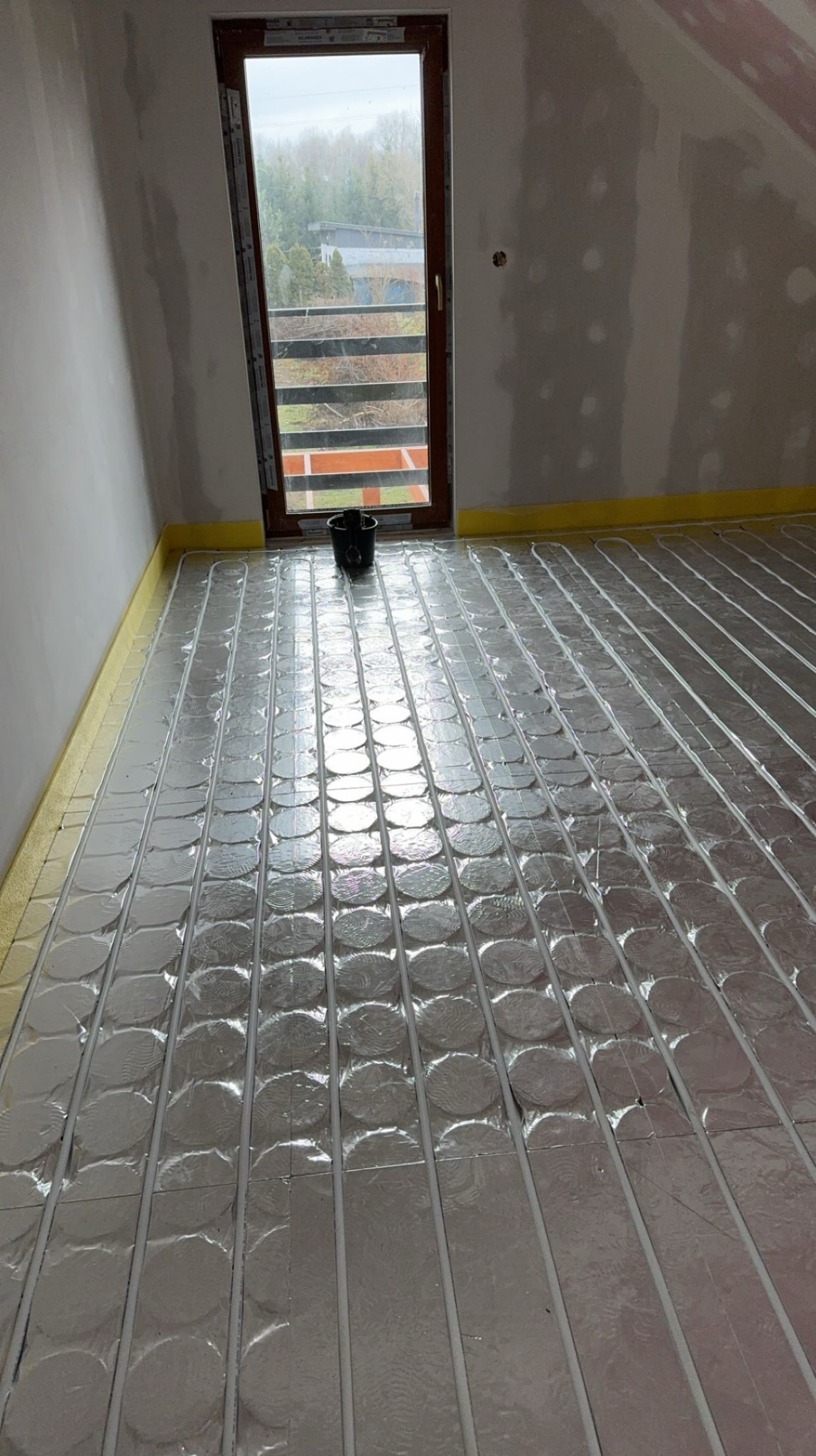 Rozłożona izolacja z folią aluminiową pod ogrzewanie podłogowe w surowym pomieszczeniu z oknem balkonowym i żółtą taśmą przy ścianach.