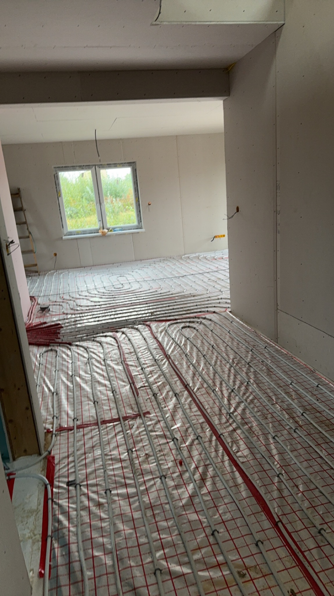 Wnętrze w trakcie montażu ogrzewania podłogowego: rury grzewcze ułożone na folii z czerwoną siatką, widok przez otwór drzwiowy, surowe ściany w tle.