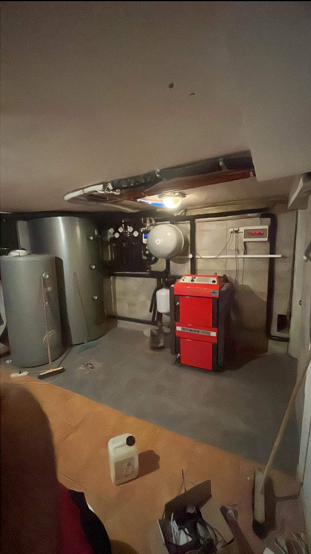 Kotłownia z czerwonym kotłem na paliwo stałe Atmos, dwoma szarymi zbiornikami akumulacyjnymi, instalacją hydrauliczną pod sufitem i kanistrem na podłodze.