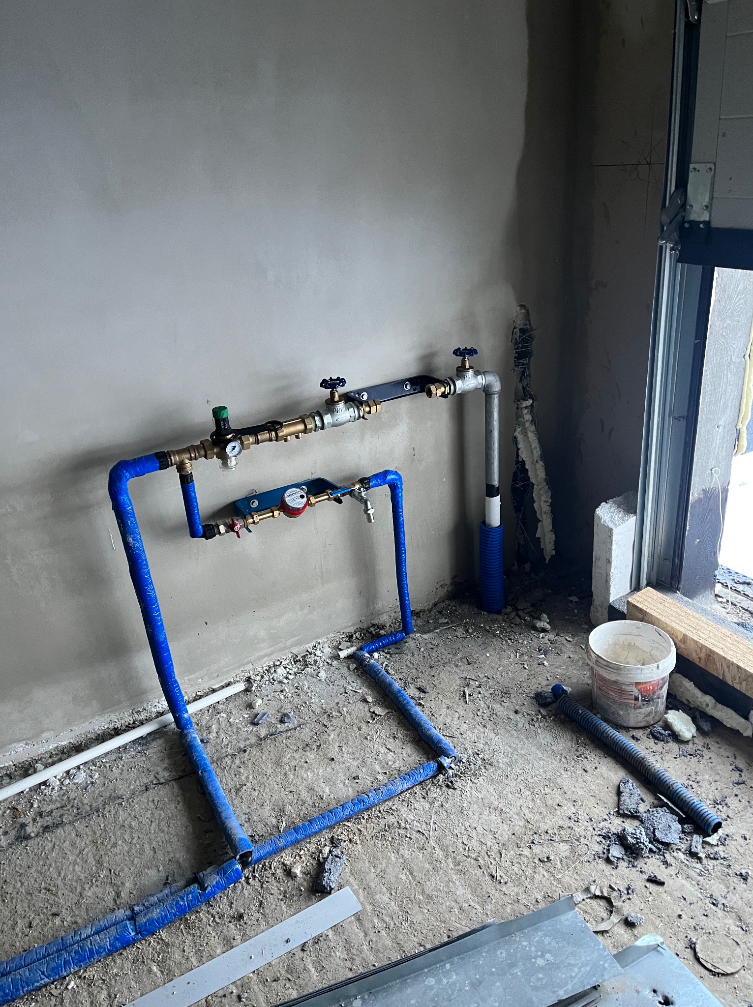 Surowa instalacja wodna z niebieskimi rurami, manometrem i zaworami, w trakcie montażu w pomieszczeniu z surowymi ścianami i betonową podłogą z resztkami gruzu.