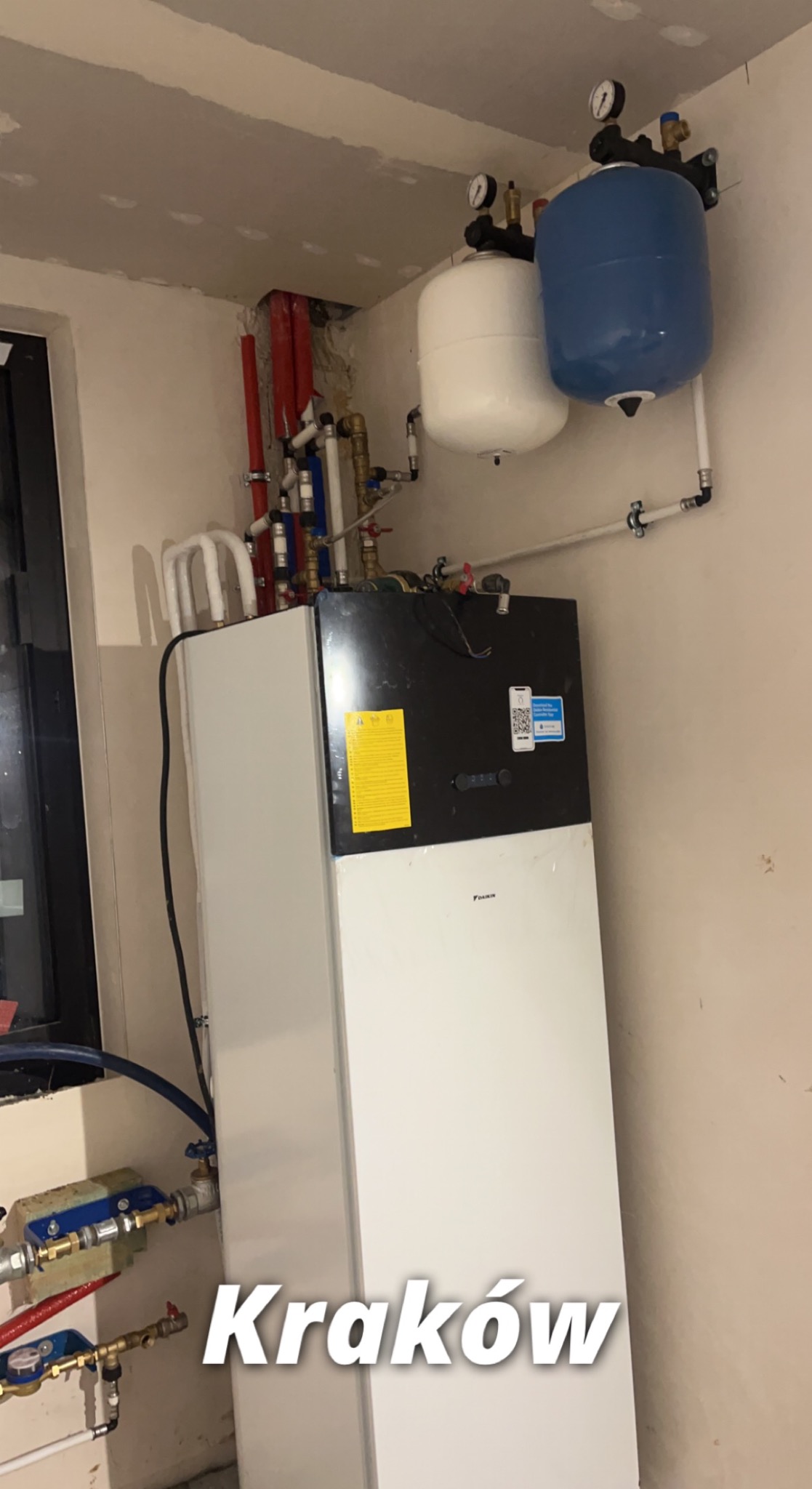 Biała pompa ciepła Daikin z podłączonymi rurami i zbiornikami wyrównawczymi w kolorze białym i niebieskim, zamontowana w pomieszczeniu z surowymi ścianami.