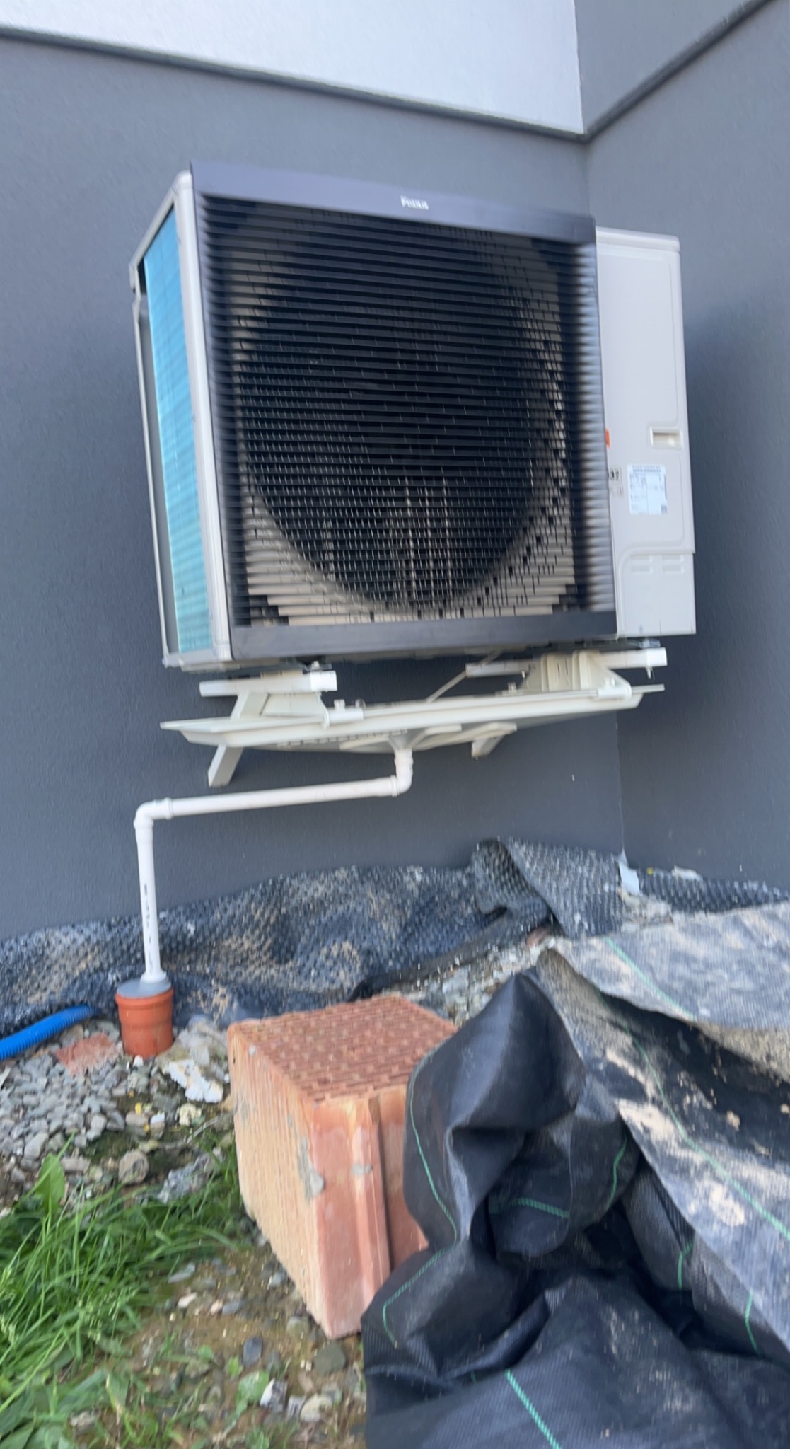 Zewnętrzna jednostka pompy ciepła zamontowana na ścianie budynku, z widocznym odprowadzeniem skroplin. W tle widoczna ściana budynku, a na pierwszym planie gruz i resztki materiałów budowlanych.