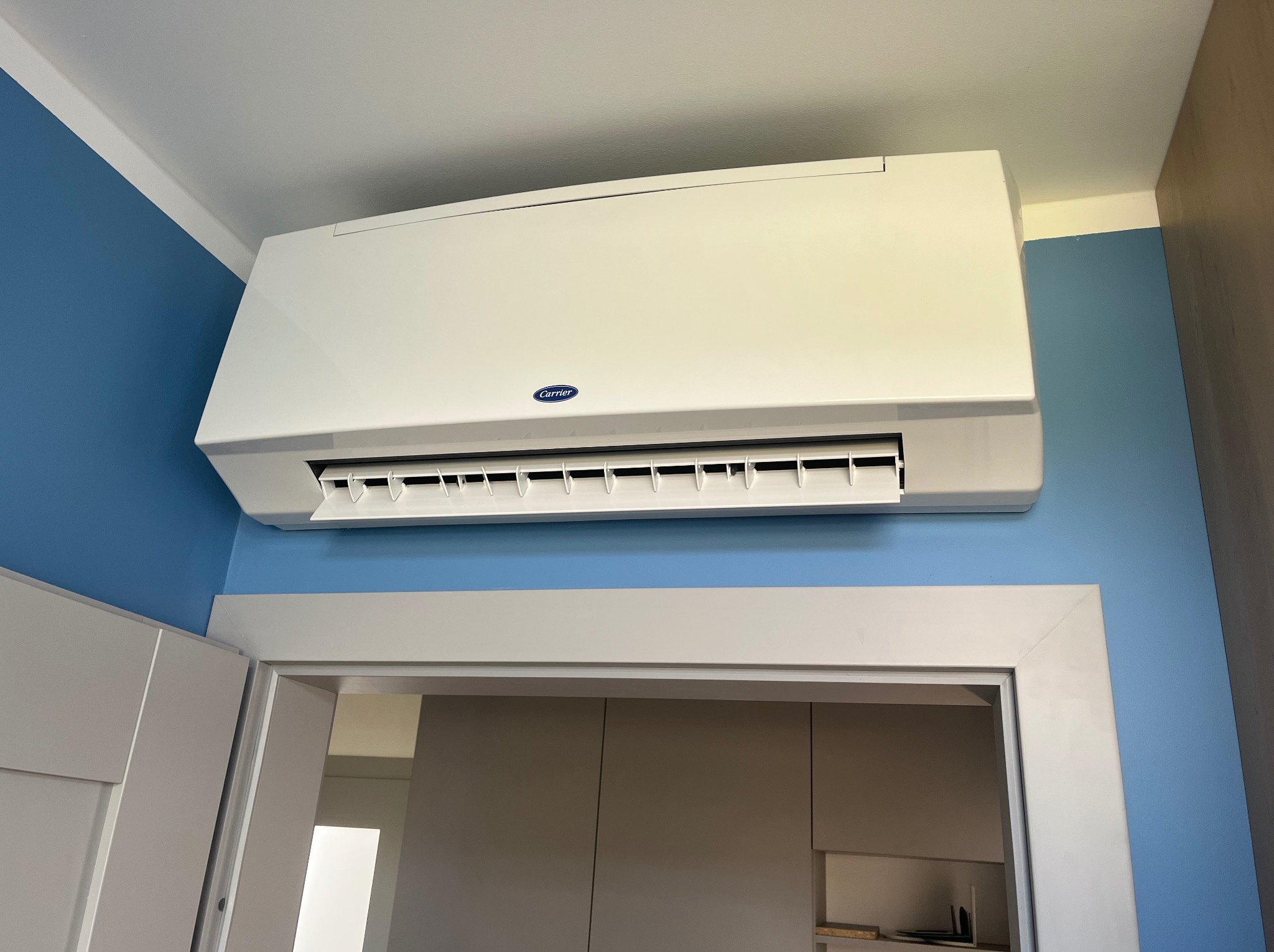 Biały klimatyzator Carrier zamontowany na błękitnej ścianie nad białymi drzwiami, widoczne żaluzje w pozycji otwartej.