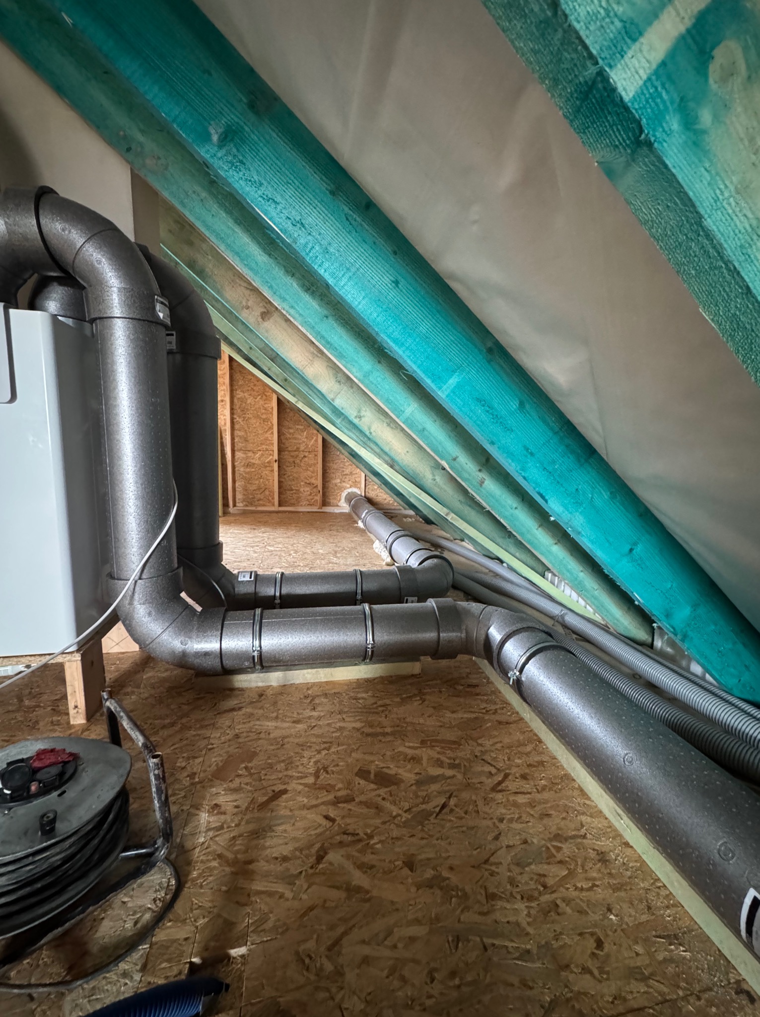 Instalacja rekuperacji w Mszanie Górnej: Widok na system rur wentylacyjnych z izolacją termiczną, poprowadzonych pod skośnym dachem w surowym stanie wykończenia poddasza.