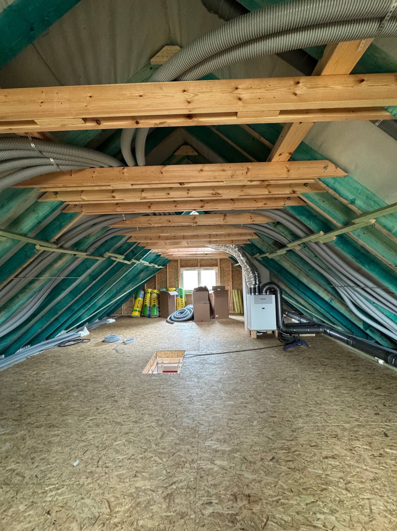 Poddasze w trakcie instalacji systemu wentylacji mechanicznej z rekuperacją, widoczne rury wentylacyjne, centrala rekuperacyjna, izolacja oraz drewniana konstrukcja dachu.