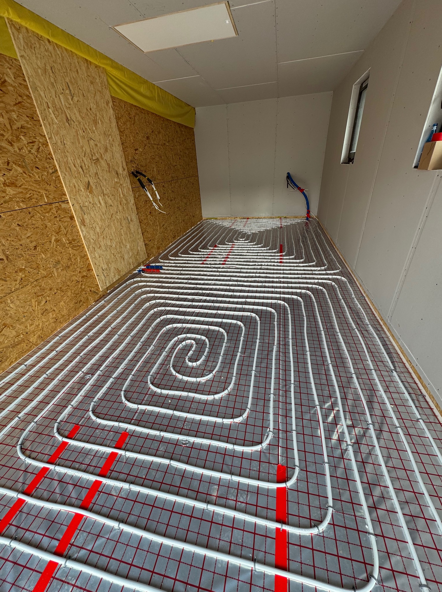 Białe rury ogrzewania podłogowego ułożone spiralnie na czerwonej siatce w nowym budynku, widoczne fragmenty ścian i okien.