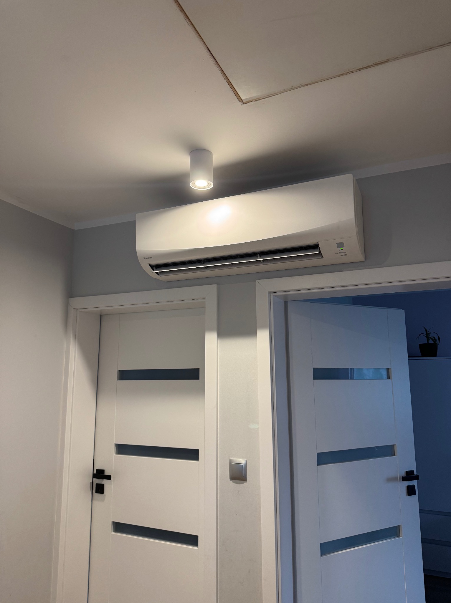 Biała klimatyzacja Daikin zamontowana nad dwoma białymi drzwiami z czarnymi klamkami w szarym pomieszczeniu, oświetlona punktowym światłem.