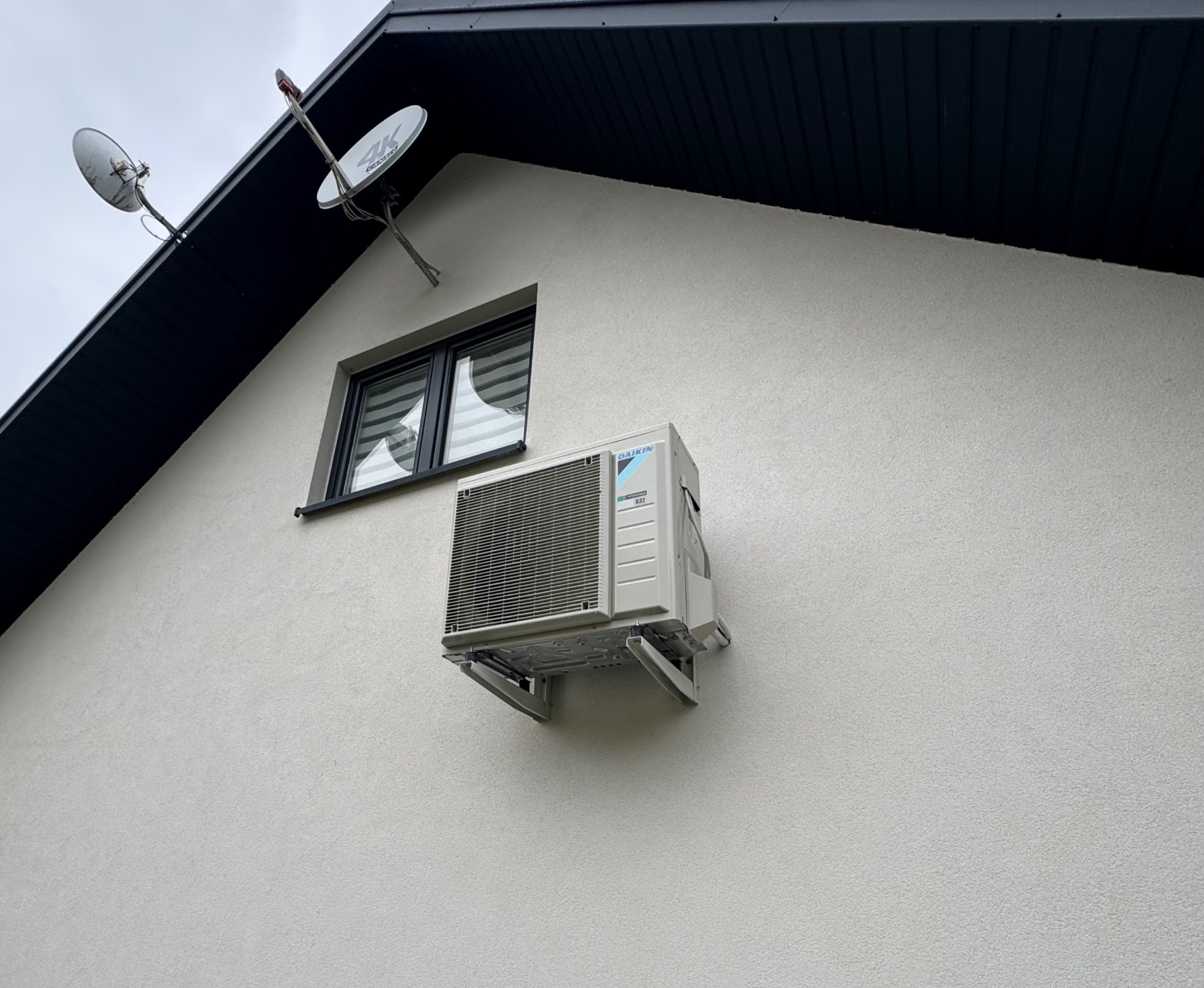 Zamontowana jednostka zewnętrzna klimatyzacji Daikin na elewacji domu z oknem i antenami satelitarnymi. Widoczne wsporniki montażowe i fragment dachu.