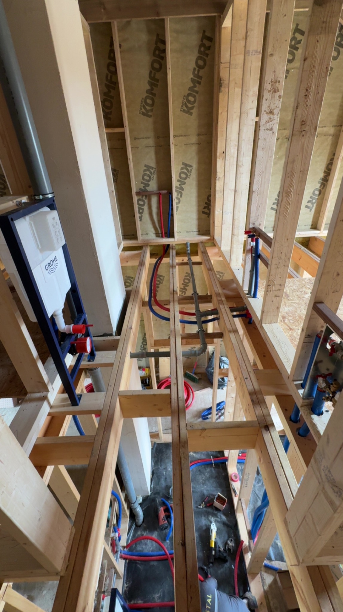 Instalacja podtynkowa WC Grohe w drewnianej konstrukcji. Widoczne rury doprowadzające wodę (czerwone i niebieskie) oraz elementy montażowe. Całość w surowym stanie, przed wykończeniem.