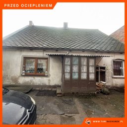 MDMS HOLDING SPÓŁKA Z OGRANICZONĄ ODPOWIEDZIALNOŚCIĄ - Stary, nieocieplony dom z szarą elewacją, widoczne zniszczenia i odpadający tynk, okno z brązową ramą, drewniane drzwi z firankami, gołębie na daszku, fragment samochodu na pierwszym planie.