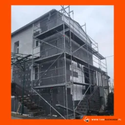 Dom w trakcie ocieplania: szara elewacja częściowo pokryta czarnym styropianem, widoczne rusztowania i fragment schodów wejściowych.