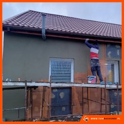 MDMS HOLDING SPÓŁKA Z OGRANICZONĄ ODPOWIEDZIALNOŚCIĄ - Pracownik na rusztowaniu, nakładający tynk na elewację domu jednorodzinnego, widoczny fragment ocieplenia, puszka z tynkiem dekoracyjnym.