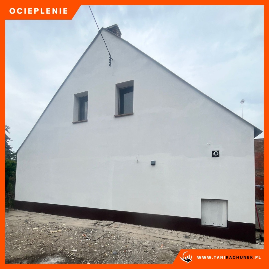 Biała elewacja domu z dwoma oknami, brązowym cokołem i niewielkim otworem wentylacyjnym w dolnej części, widoczne ślady po niedawnych pracach.