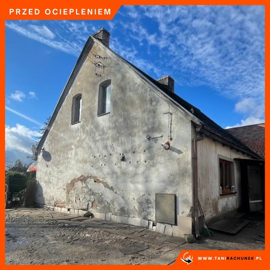 Stary, nieocieplony dom z uszkodzoną elewacją, widoczne ubytki tynku i ślady po naprawach, antena satelitarna na ścianie, zdjęcie wykonane w słoneczny dzień.