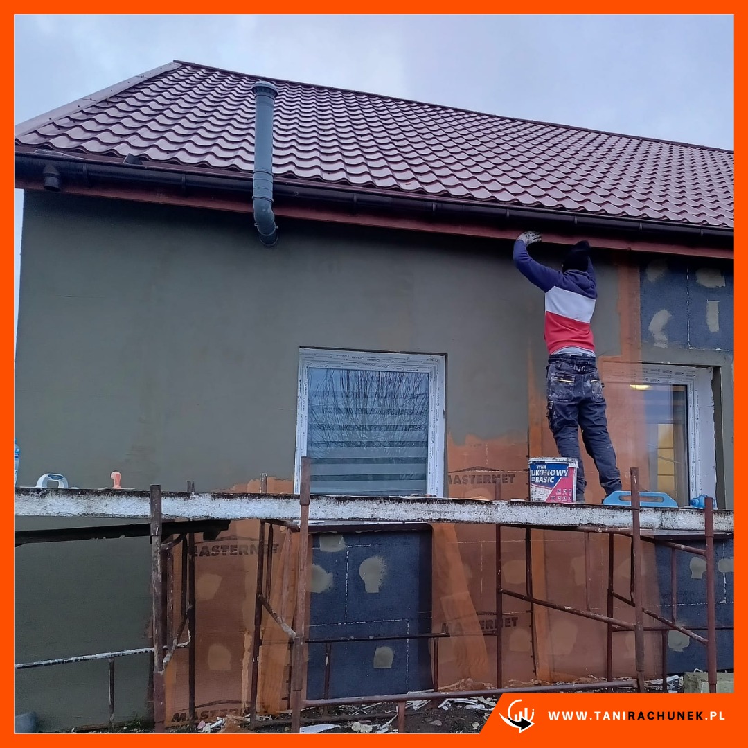 Pracownik na rusztowaniu, nakładający tynk na elewację domu jednorodzinnego, widoczny fragment ocieplenia, puszka z tynkiem dekoracyjnym.