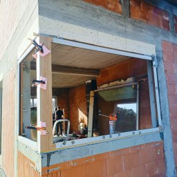 Stolarnia Hypermont - Montaż okna w surowym budynku z cegły. Widoczne klamry mocujące i pianka uszczelniająca. W tle pracownik na drabinie.