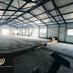 Stolarnia Hypermont - Wnętrze hali w trakcie budowy: widoczne okna, konstrukcja dachu z niebieskich belek, przygotowane podłoże. W tle pracownicy. Logo firmy w rogu.