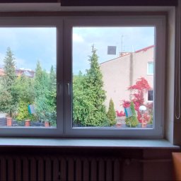 Stolarnia Hypermont - Białe okno PCV z widokiem na zieleń i budynki. Na parapecie butelka wody, obok grzejnik. Po prawej stronie zwisająca roślina doniczkowa.