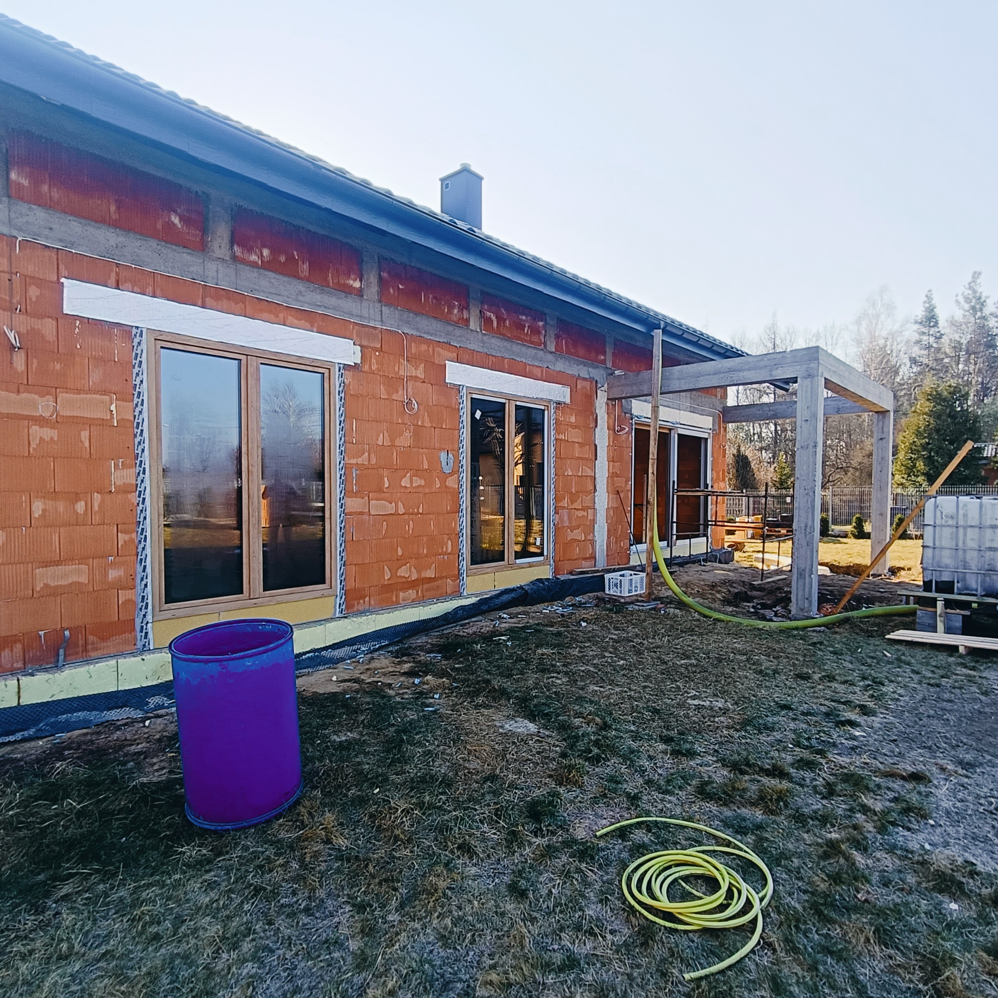 Budowa domu z oknami w stanie surowym otwartym, widoczny fragment elewacji z cegły, drewniana konstrukcja zadaszenia tarasu i fioletowa beczka na trawniku.