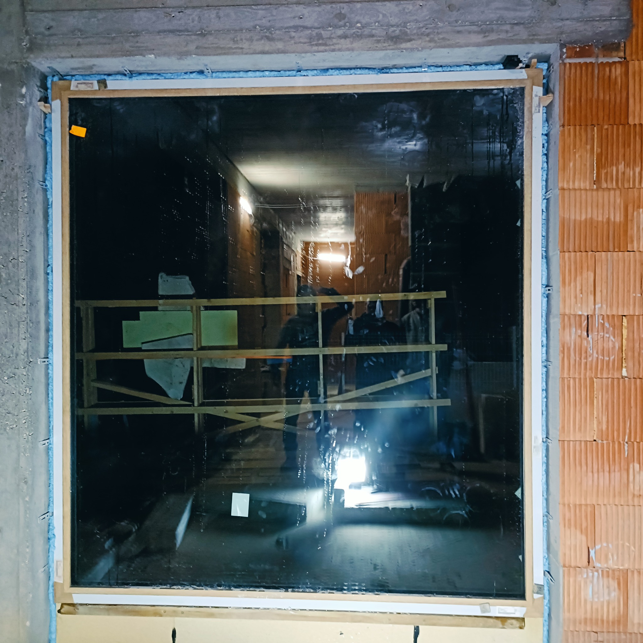 Montaż okna PCV w surowym stanie. Widoczne pianka montażowa, cegły i beton. W tle rusztowanie i ekipa montażowa. Okno zabezpieczone folią.