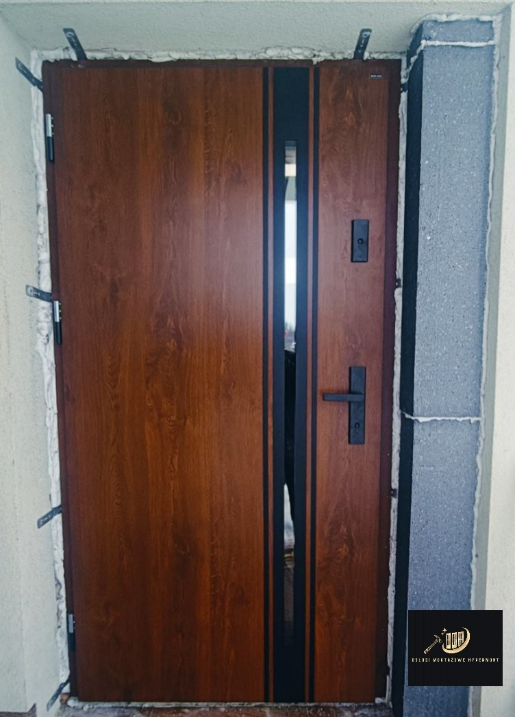 Zamknięte, brązowe drzwi wejściowe z czarnymi paskami i szybą, osadzone w ościeżnicy z widoczną pianką montażową i elementami mocującymi.